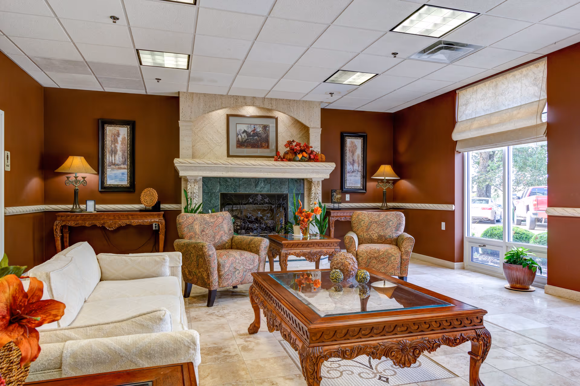 A cozy living room area with a white sofa, two patterned armchairs, and two ornately carved wooden coffee tables with glass tops. The room features a decorative fireplace with a framed picture above it, two table lamps on side tables, and warm brown walls. A large window with a beige roman shade lets in natural light and shows a view of parked cars and greenery outside.