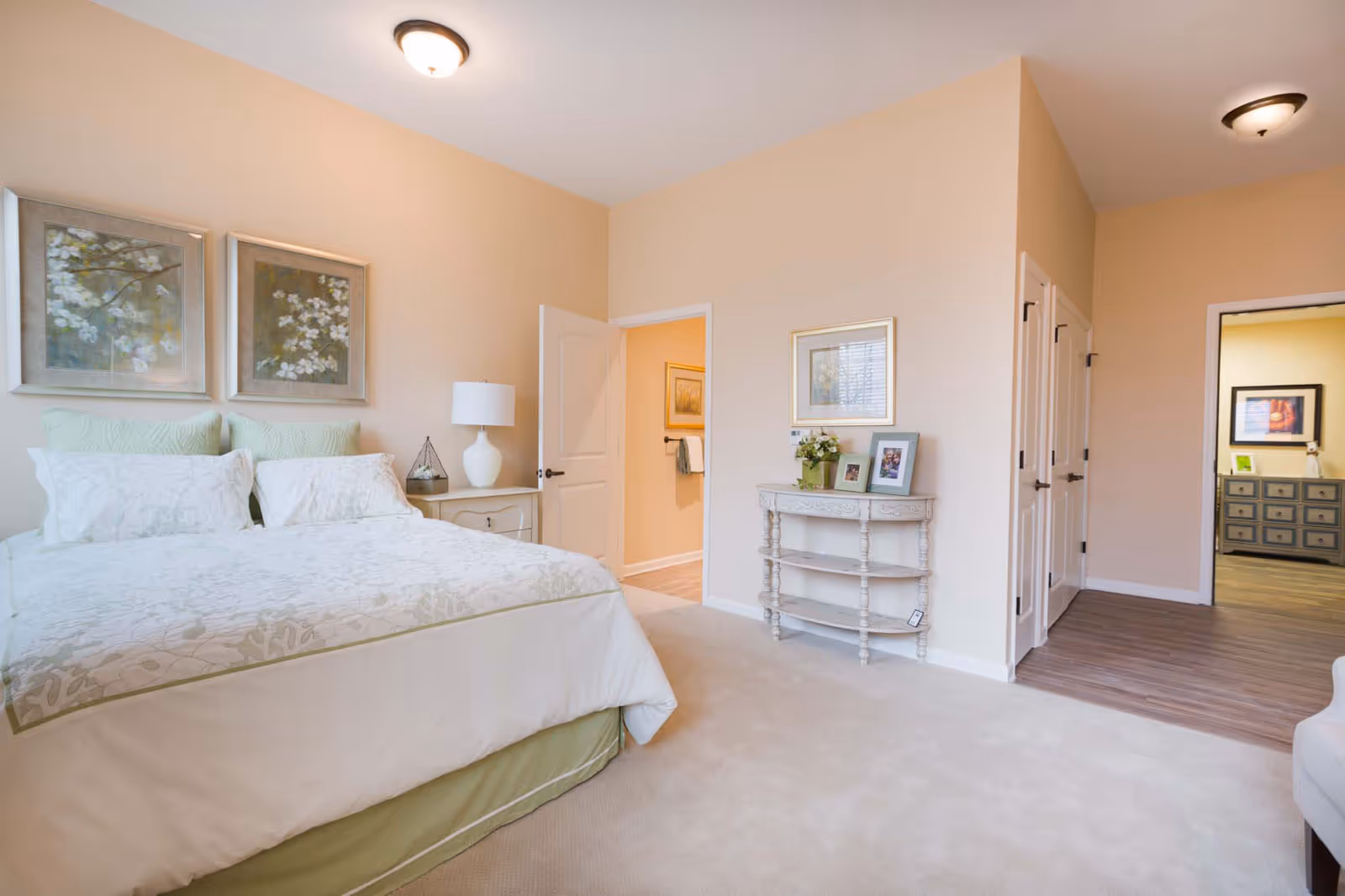 Bright furnished bedroom with a made bed, nightstand and lamp, decorative artwork, and an open doorway to a hallway and adjacent room.