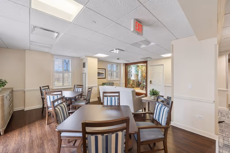 Interior view of a senior living facility common area with wooden tables and chairs featuring striped cushions, a white couch, a side table with a plant, windows with white shutters, and an exit sign on the ceiling.