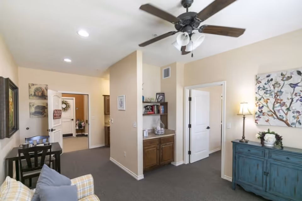 Interior view of a living area in an assisted living facility featuring a ceiling fan with lights, a small wooden table with chairs, a blue sideboard with a lamp and floral decoration, wall art, and an open doorway leading to other rooms.