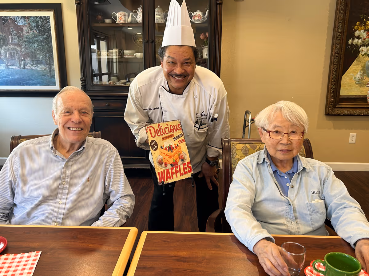 A smiling chef stands between two seated elderly residents at a dining table, holding a sign that reads 'Delicious Waffles'.