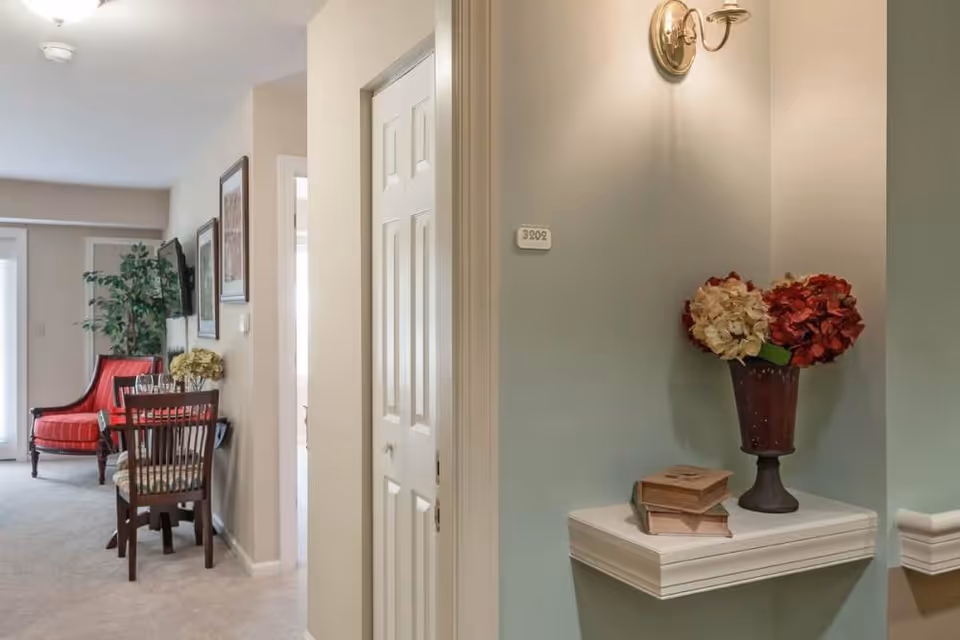 Interior view of a senior living facility room at Chancellor's Village showing a small corner shelf with a vase of red and white flowers and two books. The room has beige walls, a wall-mounted light fixture, a white door, a dining table with chairs, a red upholstered chair, framed pictures on the wall, and a potted plant near a window with blinds.