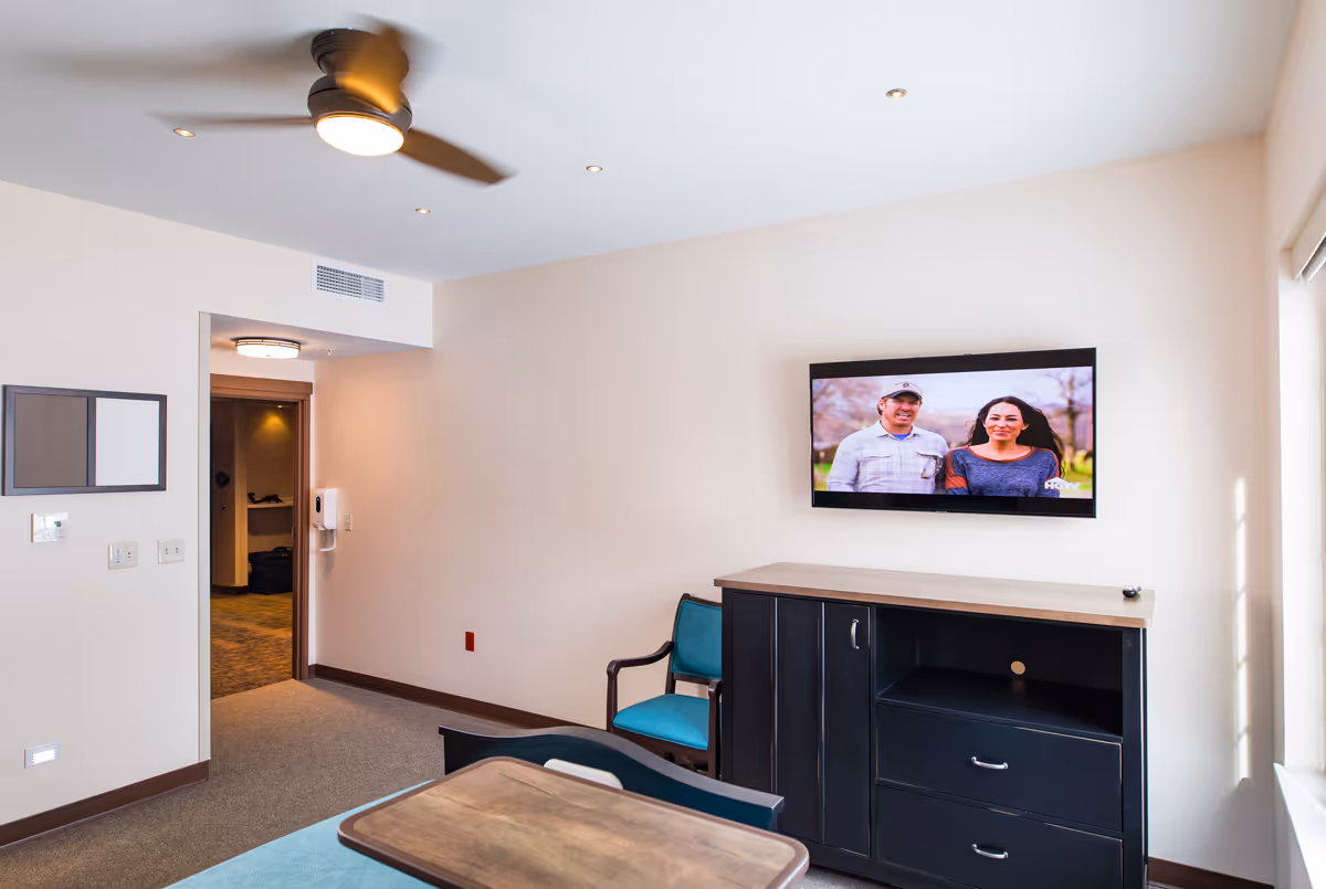 A bright senior living room with a ceiling fan, a wall-mounted flat screen TV showing a man and woman, a black cabinet with drawers, a teal chair, and a wooden table in the foreground. The room has beige walls and carpeted floor, with an open doorway leading to another area.