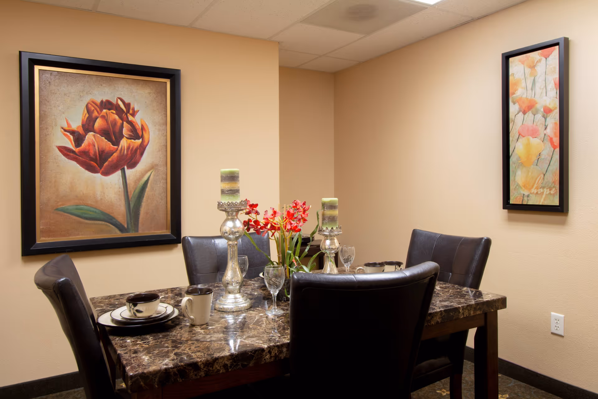 A dining area with a dark marble table set with plates, cups, and glasses. Four black leather chairs surround the table. Two silver candle holders with green candles and a vase with red flowers are placed on the table. The beige walls have two framed floral paintings.