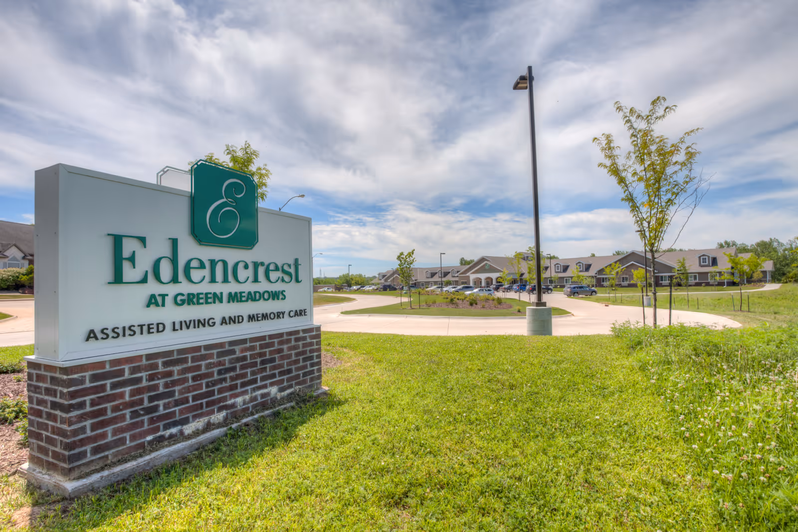 Outdoor view of Edencrest at Green Meadows assisted living and memory care facility sign with the building and parking lot in the background under a partly cloudy sky.