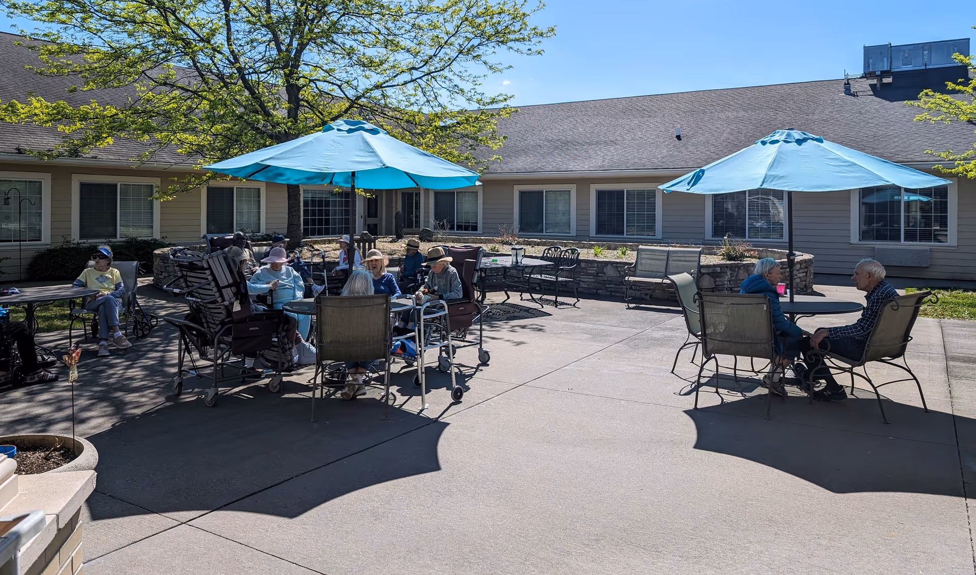 Outdoor patio area at Rock Run Memory Care with elderly residents sitting around tables under blue umbrellas. The patio is paved and surrounded by a single-story building with windows. Trees provide some shade and greenery.