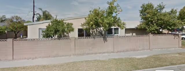 Single-story beige facility behind a low wall with trees and a sidewalk in front.