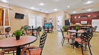A spacious common area with multiple round tables and chairs with red patterned cushions. The room has beige walls, a mounted flat-screen TV, windows with blinds, and a door leading outside. There are plants on the tables and a kitchenette area with dark wood cabinets, a microwave, and a refrigerator in the background.