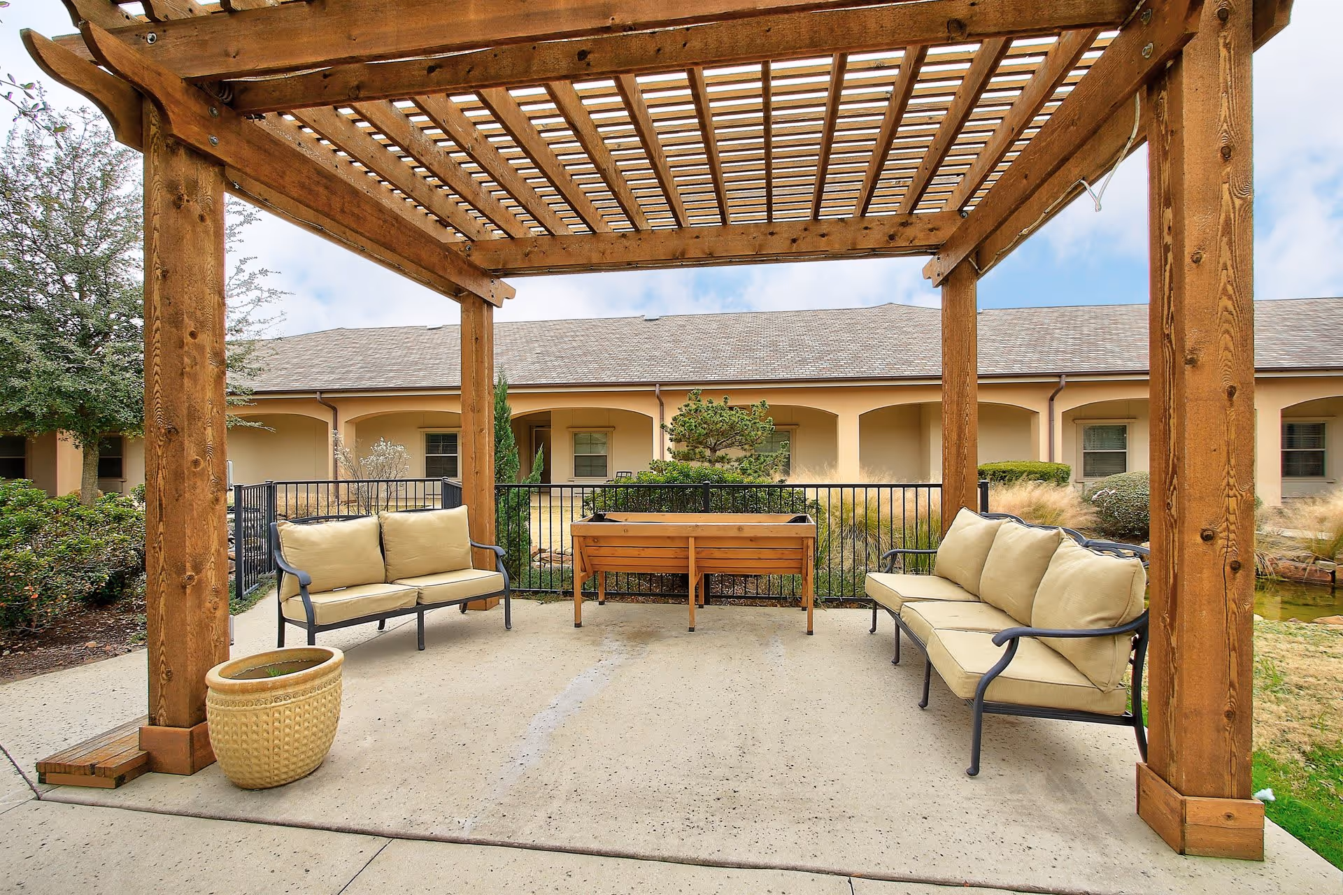 Outdoor seating area with a wooden pergola overhead, two cushioned benches facing each other, a planter pot, and a raised garden bed in the background, surrounded by greenery and a building with arched windows.