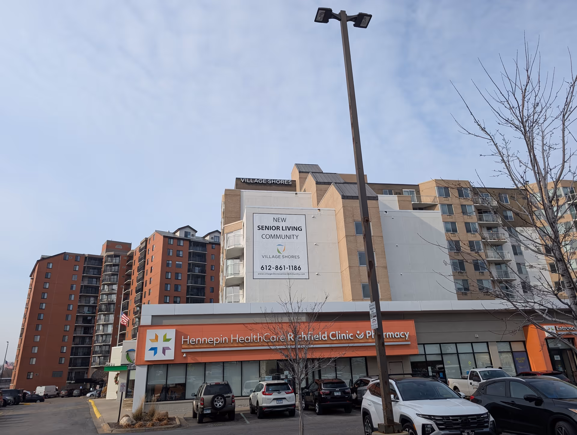 Exterior view of a multi-story building with a sign that reads 'Village Shores' at the top. Below, there is a large banner advertising a new senior living community with contact information. In front of the building is a Hennepin HealthCare Richfield Clinic & Pharmacy with several parked cars and a tall street lamp. Leafless trees are visible in the parking lot under a partly cloudy sky.