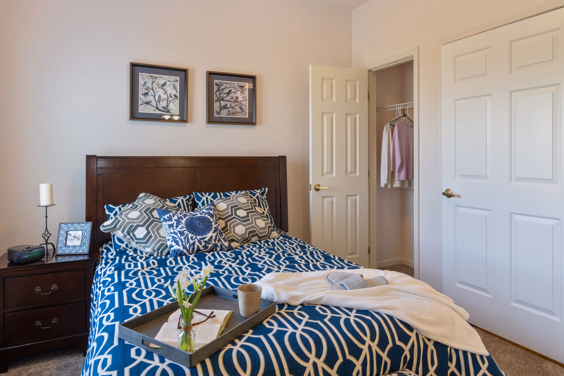 A bedroom with a wooden headboard bed covered in blue and white patterned bedding and multiple pillows. A tray with a vase of flowers, a book, a cup, and a white cloth is placed on the bed. There is a nightstand with a candle, a framed photo, and an alarm clock. Two framed bird pictures hang on the wall above the bed. An open door reveals a closet with clothes hanging inside.