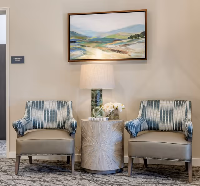 Two upholstered armchairs flank a round side table with a lamp and flowers beneath a landscape painting on a neutral wall.