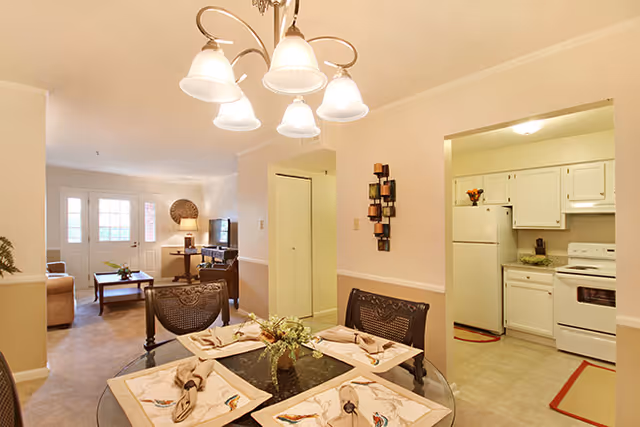 Dining area with a round glass table set for four under a chandelier, open to a living room and a white kitchen.