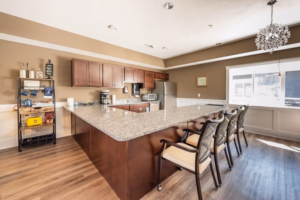 A bright kitchen area with a large granite countertop island surrounded by four chairs. The kitchen features wooden cabinets, a stainless steel refrigerator, a coffee maker, a microwave, and a small shelving unit with various kitchen items. A decorative chandelier hangs from the ceiling, and a large window lets in natural light.