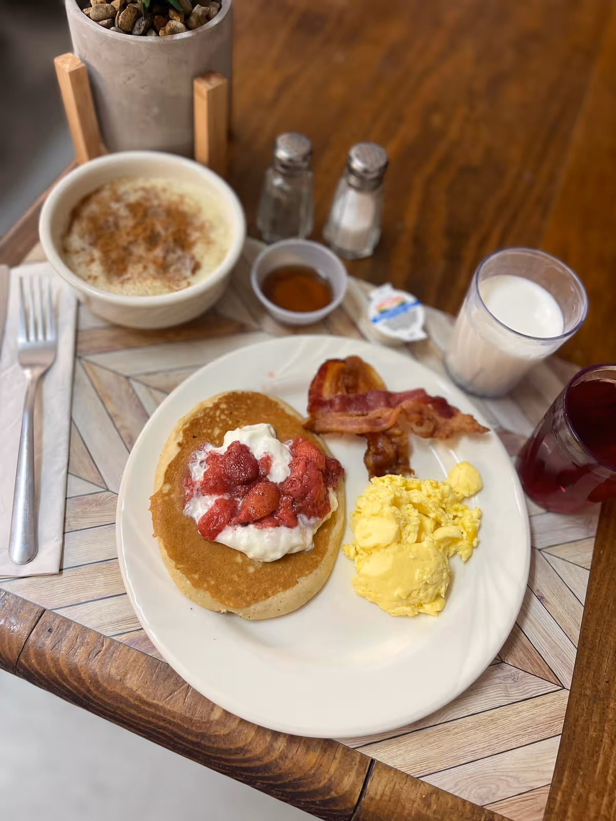 A breakfast plate on a wooden table with a pancake topped with strawberries and cream, scrambled eggs, bacon, a bowl of oatmeal, and glasses of milk and juice.
