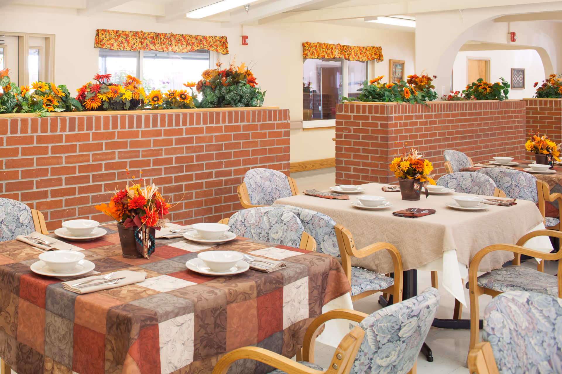 A dining area in a senior living facility featuring tables set with dishes and decorative floral arrangements.