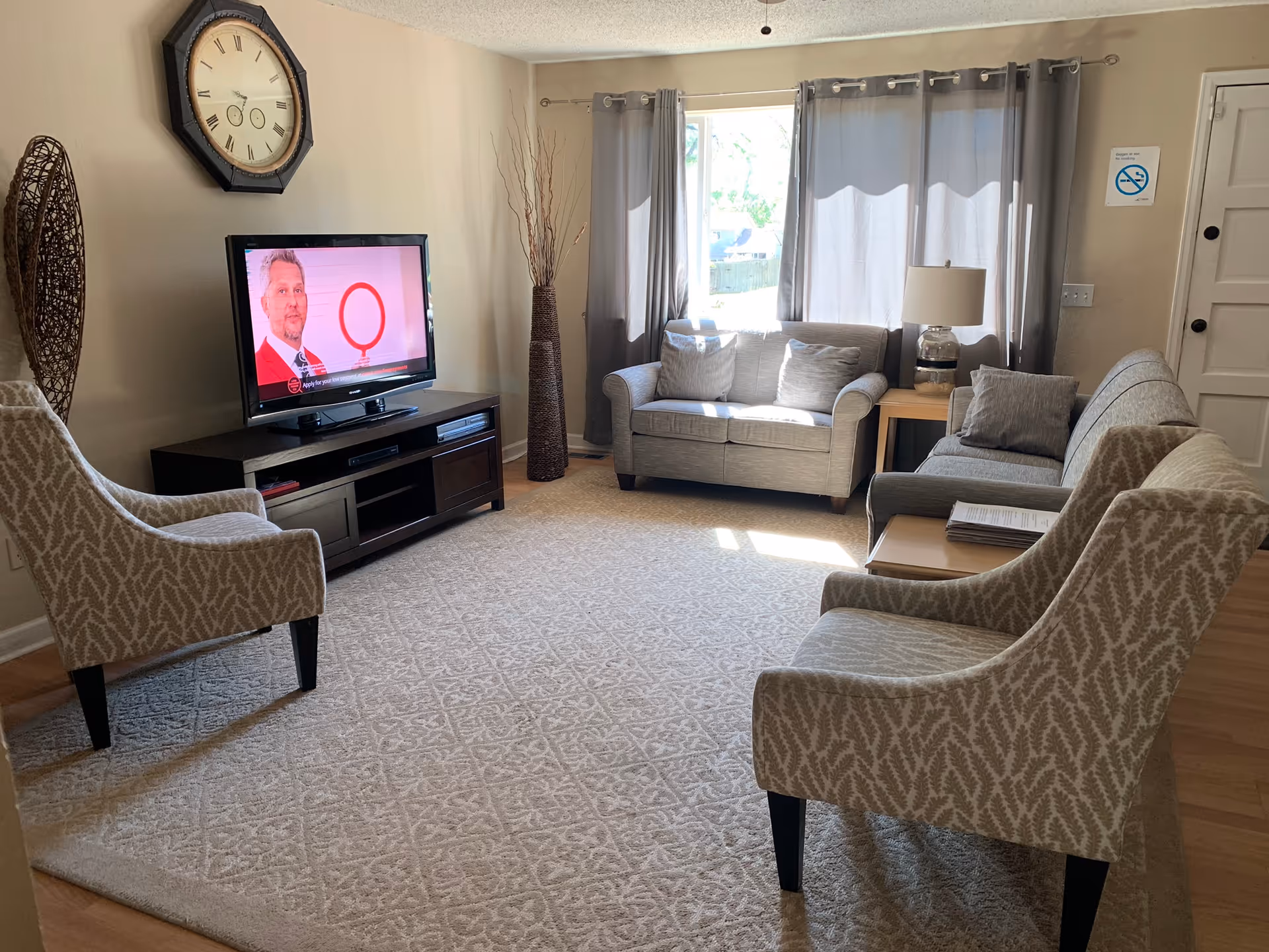 A cozy living room with two patterned armchairs, two gray sofas, a wooden TV stand with a flat-screen TV, a large wall clock, a tall decorative vase with branches, a side table with a lamp, and windows with gray curtains letting in natural light.