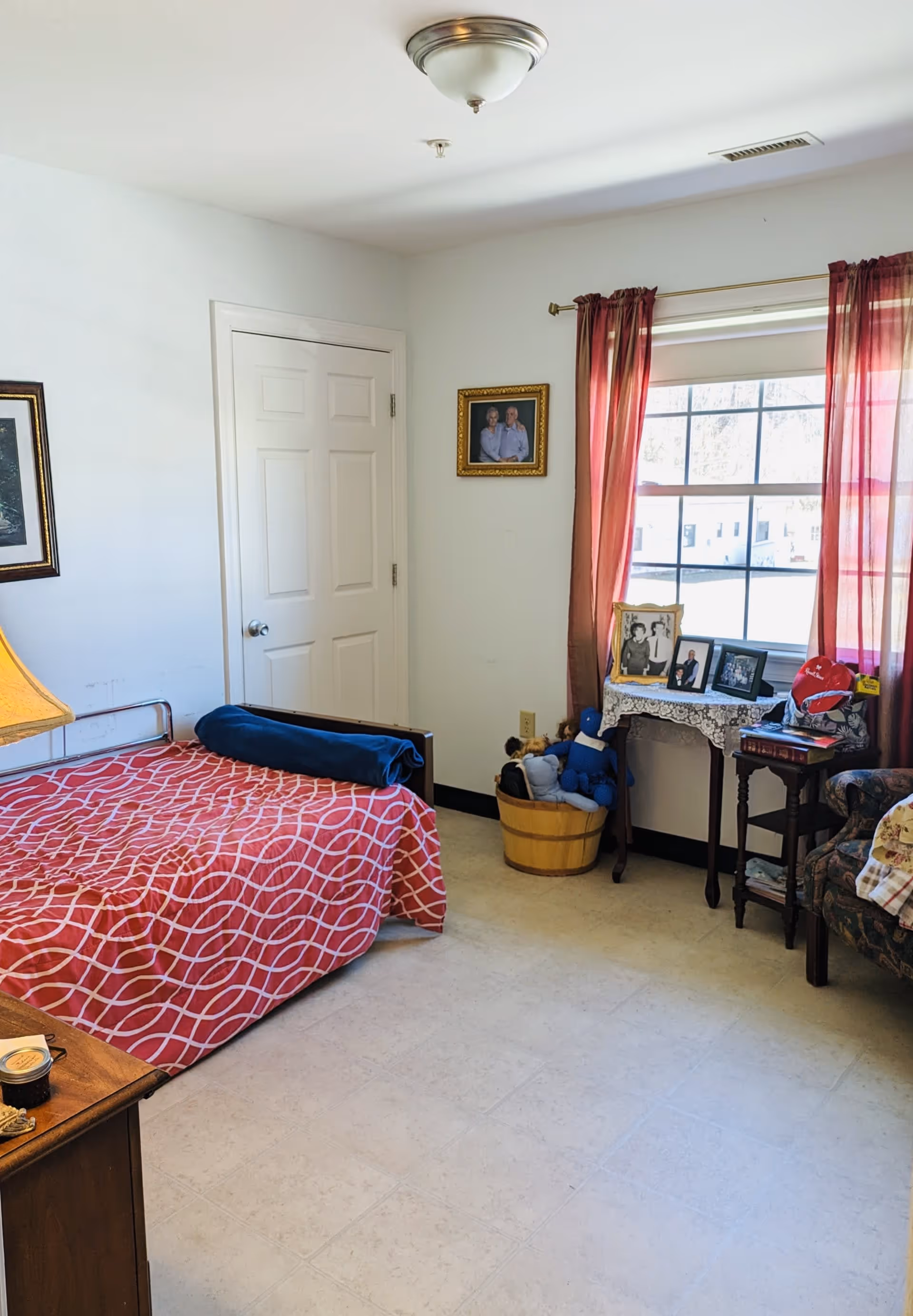 A cozy bedroom with a single bed covered in a red and white patterned blanket. There is a wooden nightstand with a lamp on the left side of the bed. A door is visible on the far wall. A window with red curtains lets in natural light, and beneath it is a small table with framed photos and a basket filled with stuffed toys. A floral armchair is partially visible on the right side.