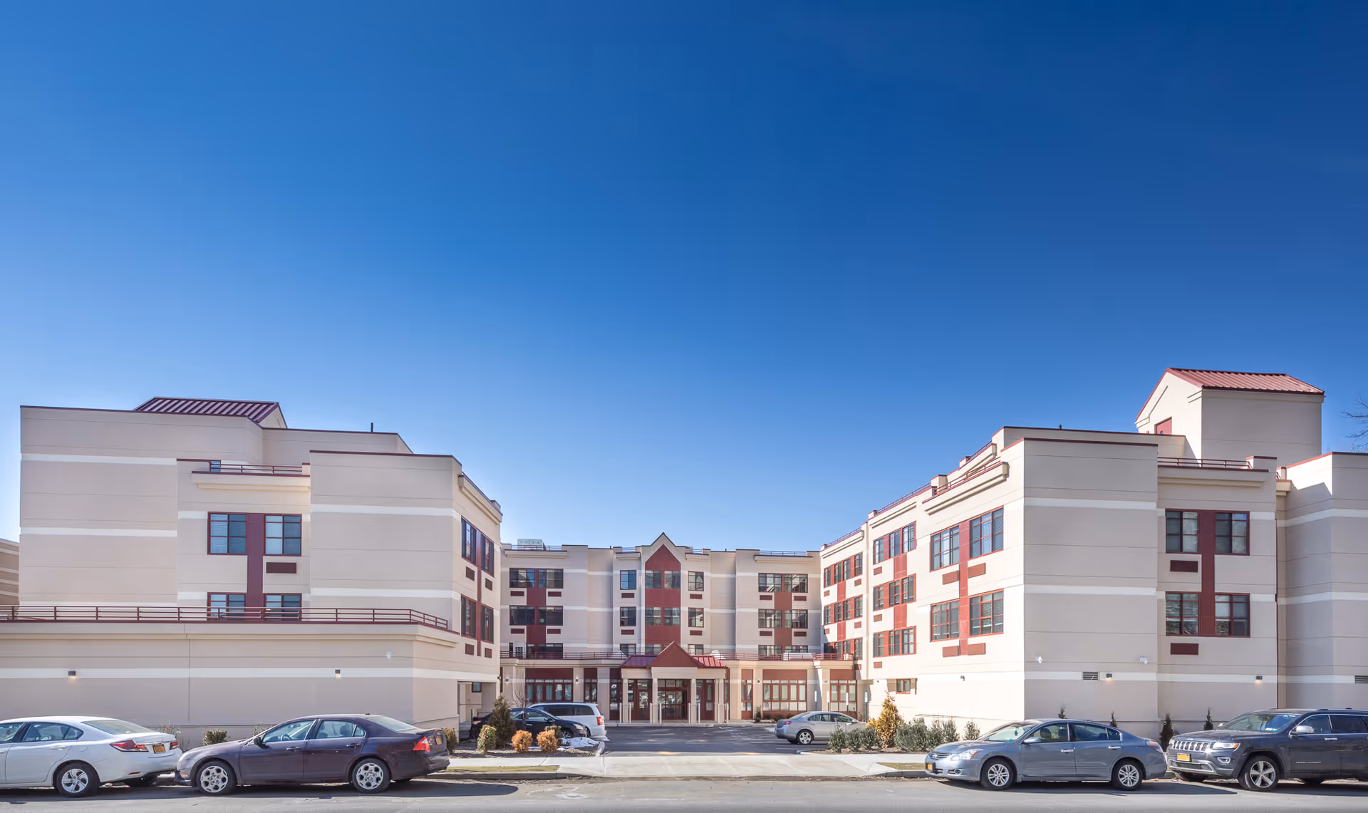 Exterior front view of a multi-story senior living facility building with beige walls and red accents under a clear blue sky. Several cars are parked in front of the building.