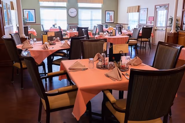 A dining room with multiple tables covered in pink tablecloths, each set with folded napkins, glasses, and condiments. The room has large windows with blinds, framed artwork on the walls, and a clock above the windows. Chairs with armrests surround the tables, and the floor is wooden.