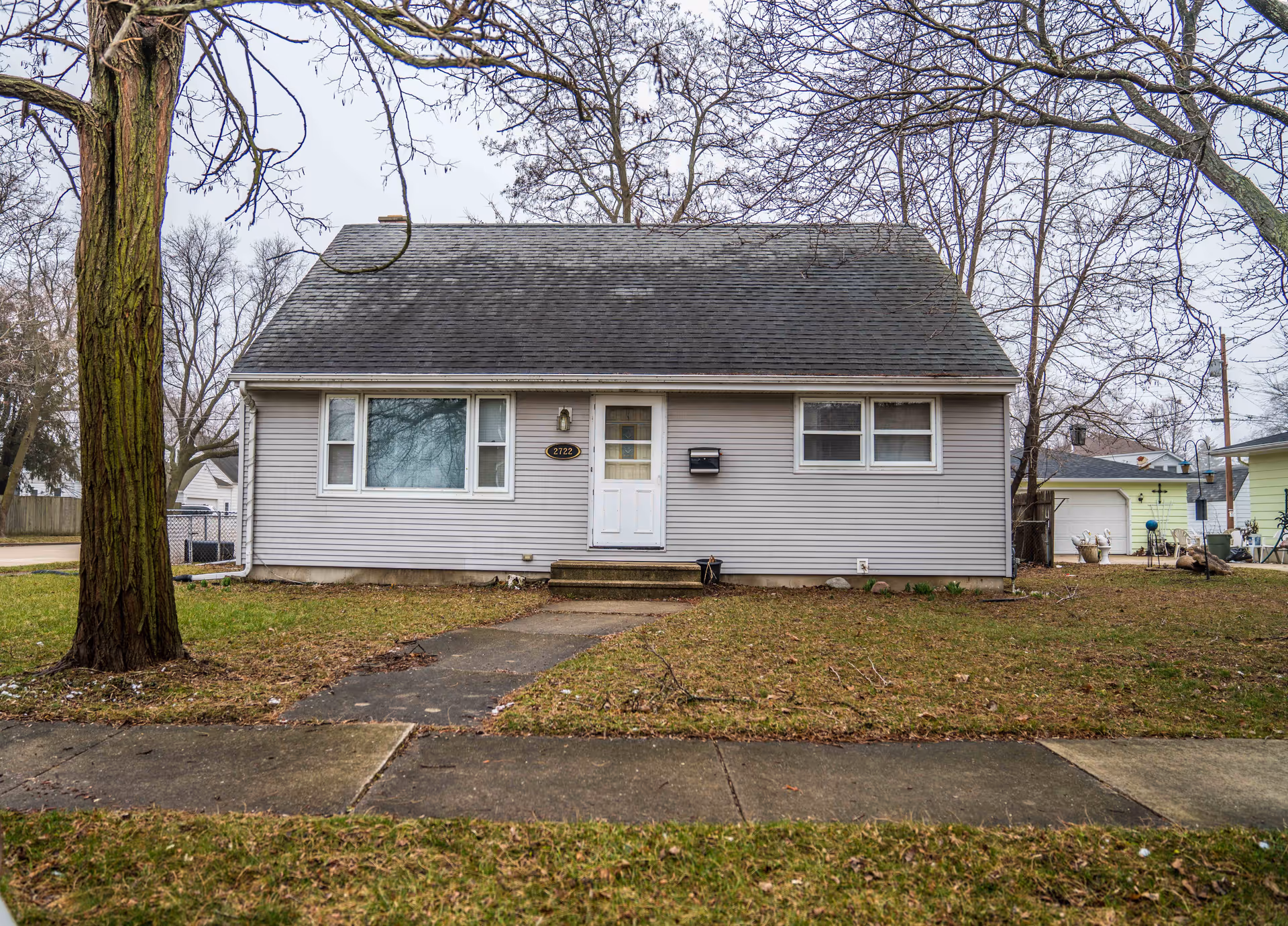Front exterior view of a small, single-story house with gray siding, a dark shingled roof, a white front door with a window, and two sets of windows on either side of the door. There is a concrete pathway leading to the front steps, a mailbox mounted next to the door, and a large tree on the left side of the lawn. The grass is patchy and there are leafless trees in the background under an overcast sky.