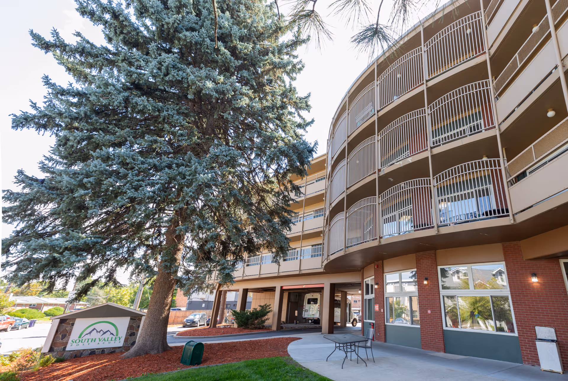 Front entrance of a multi-story curved senior living building with balconies, a large pine tree, outdoor seating, and a 'South Valley' sign.
