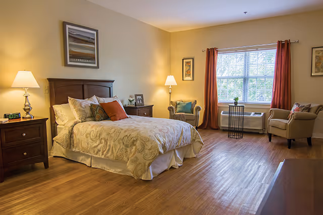 Spacious furnished bedroom with a bed, bedside tables and lamps, two armchairs by a window, and hardwood floors.