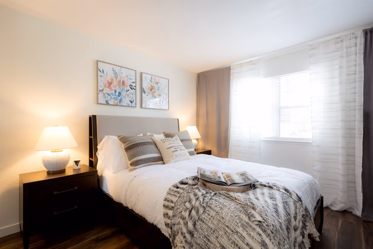 A bright bedroom with a large bed covered in white bedding and multiple pillows, a gray and white patterned throw blanket at the foot of the bed, two bedside tables each with a lamp, floral artwork on the wall above the bed, and a window with sheer and solid curtains allowing natural light to fill the room.