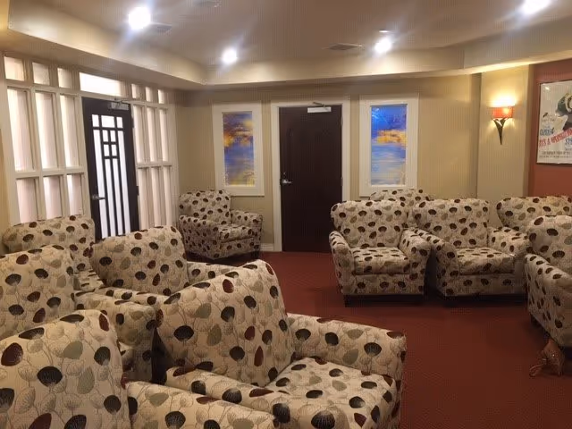 A cozy common area with multiple cushioned armchairs upholstered in a patterned fabric featuring leaves in various colors. The room has beige walls, red carpet flooring, two framed landscape paintings on the back wall, a dark wooden door in the center, and a wall sconce with a red shade on the right side. There is also a framed poster on the right wall and a decorative window with vertical slats on the left side.