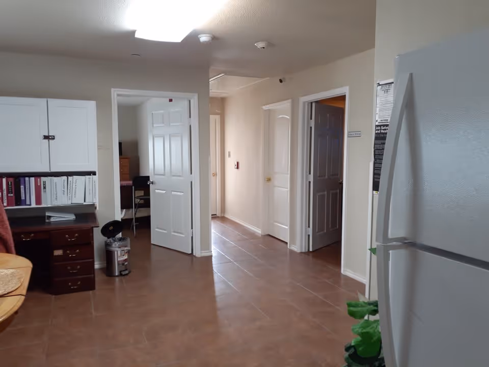 Interior view of a hallway in an assisted living facility with several open doors leading to different rooms. On the left side, there is a desk with binders and a trash can nearby. On the right side, a white refrigerator is partially visible with a plant in front of it. The floor is tiled and the walls are painted light beige.
