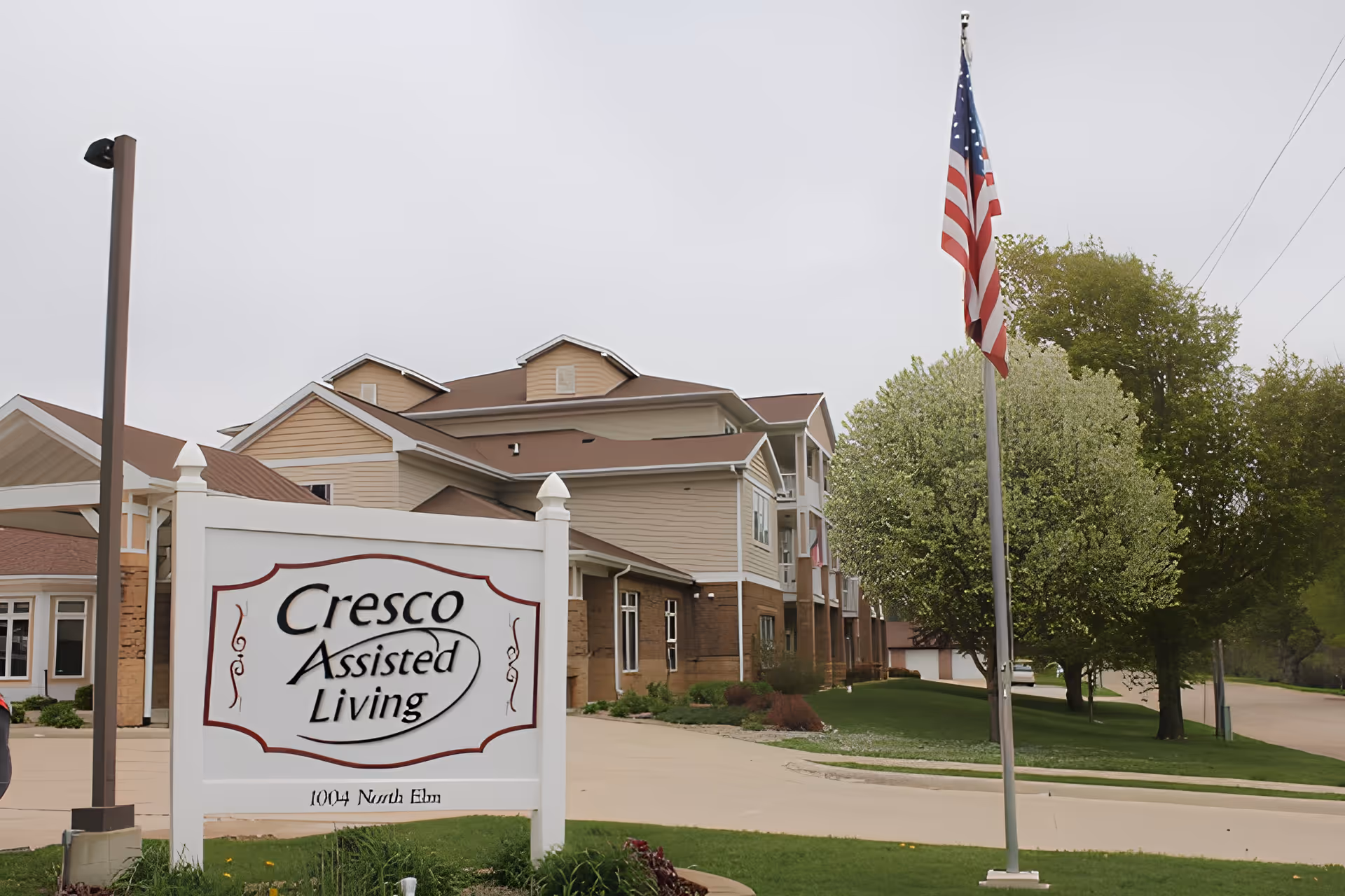 Front exterior of Cresco Assisted Living with a large entrance sign, American flag, and the multi-story building behind it.