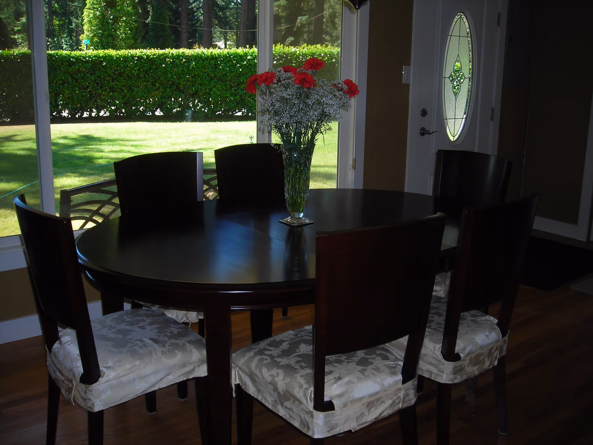 A dark wooden round dining table with six chairs that have white patterned cushions. A glass vase with red and white flowers is placed in the center of the table. Large windows behind the table show a green lawn and bushes outside. A white door with an oval decorative glass panel is visible to the right.