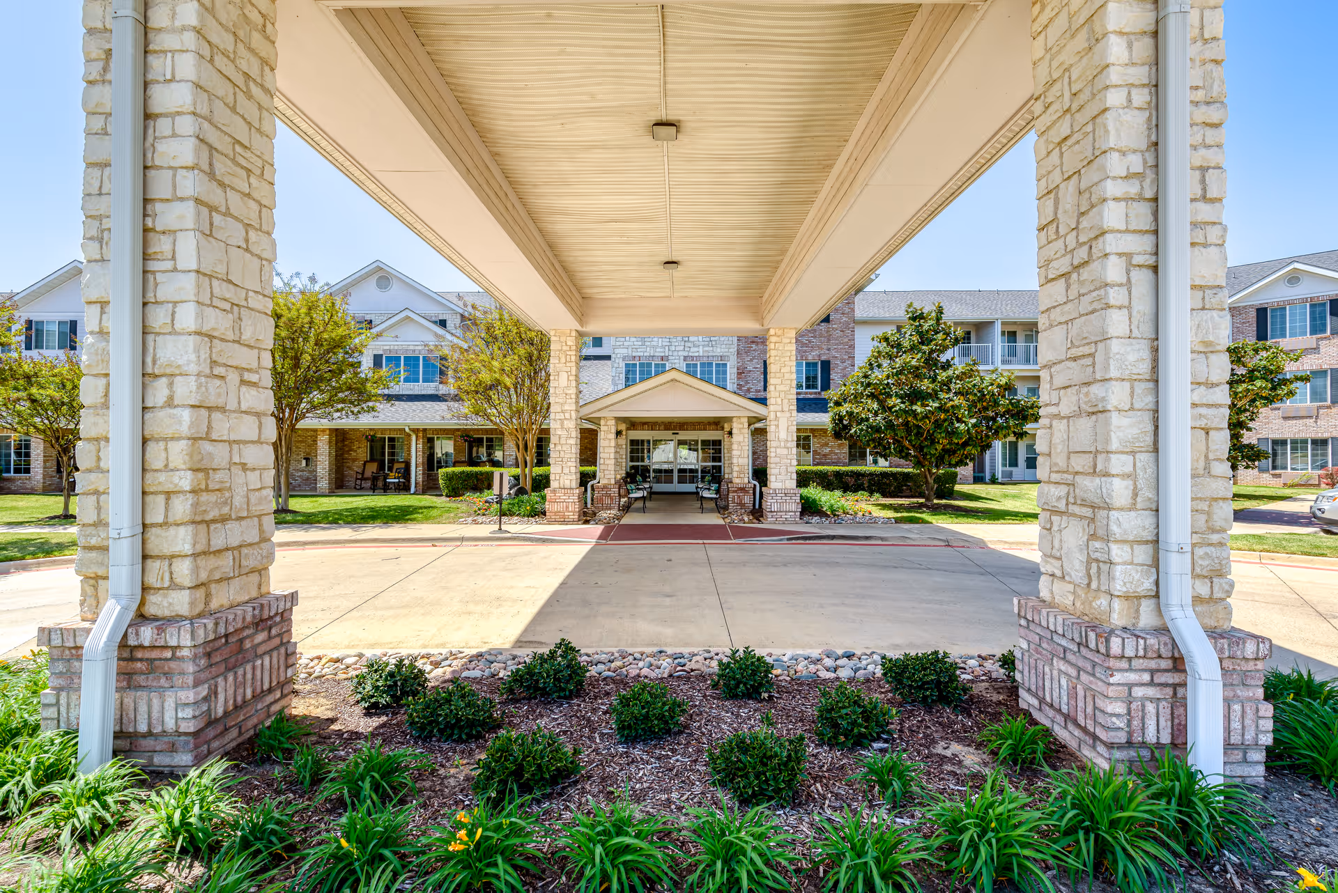 Covered porte-cochère entrance with stone columns and landscaping in front of a multi-story senior living building.