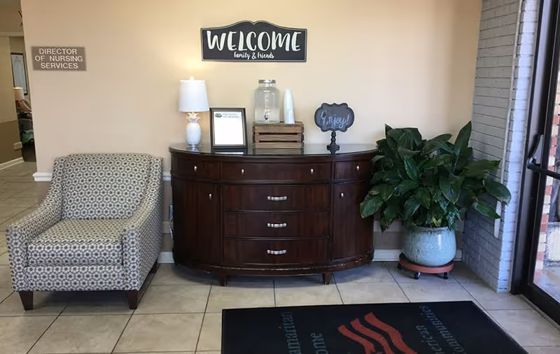 A welcoming corner inside a facility with a patterned armchair on the left, a dark wooden sideboard in the center holding a lamp, a water dispenser, and a small sign that says 'Enjoy!'. Above the sideboard is a wall sign that reads 'WELCOME family & friends'. To the right is a large potted plant near a glass door entrance. A sign on the left wall reads 'DIRECTOR OF NURSING SERVICES'.