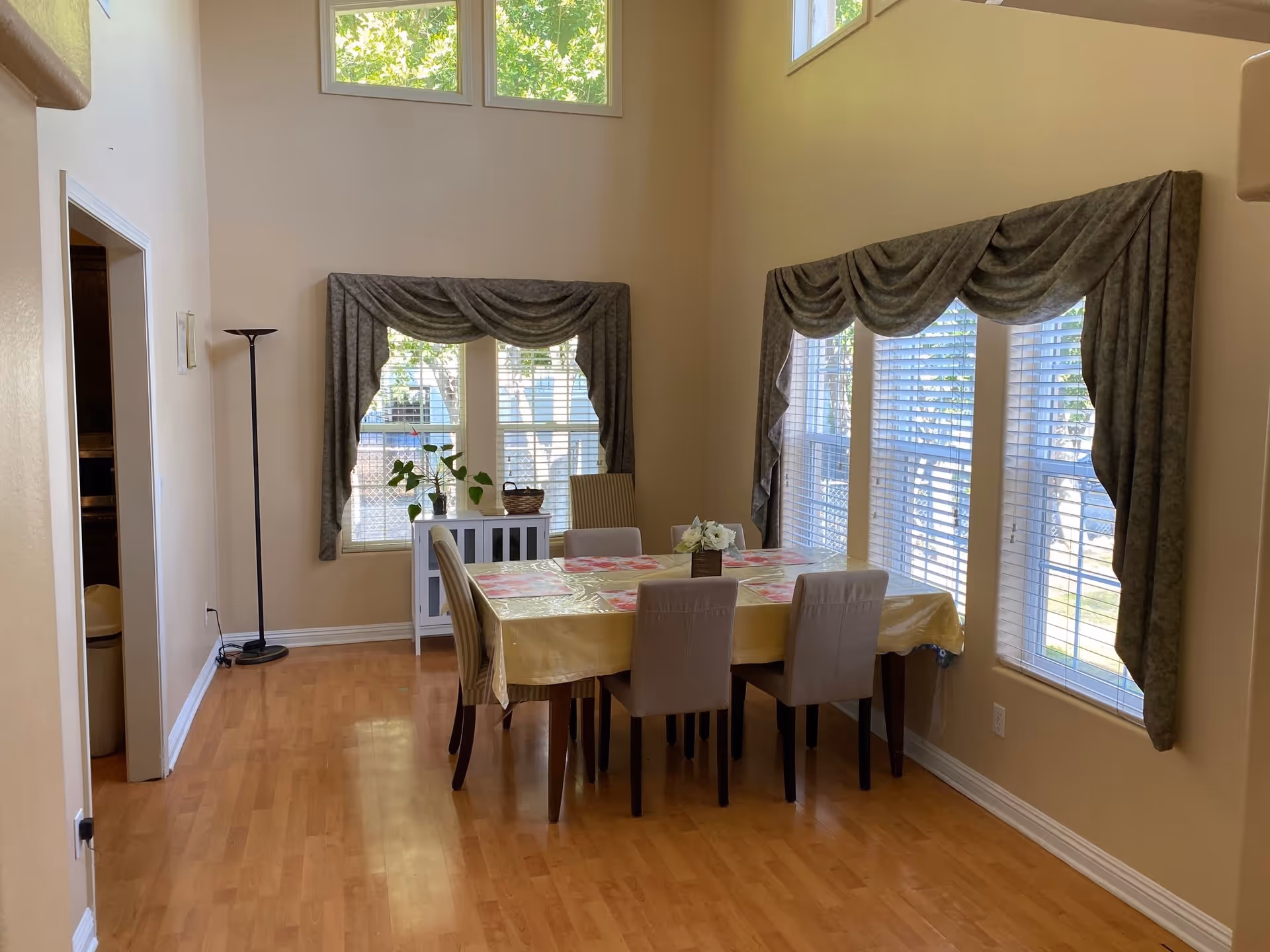 A dining room with a rectangular table covered with a yellow tablecloth and a floral centerpiece. Six chairs surround the table. The room has large windows with green draped curtains and white blinds, allowing natural light to fill the space. There is a small white cabinet with plants on top against the wall and a tall floor lamp in the corner. The floor is wooden, and the walls are painted beige.