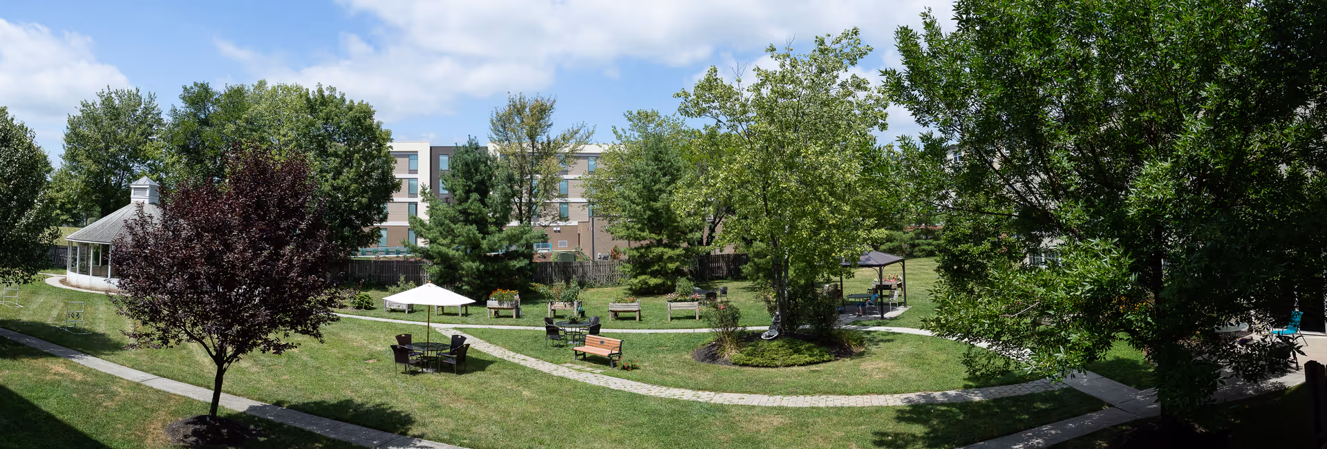 Sunny landscaped courtyard with winding brick paths, benches, umbrellas, a gazebo, and trees in front of the facility building.