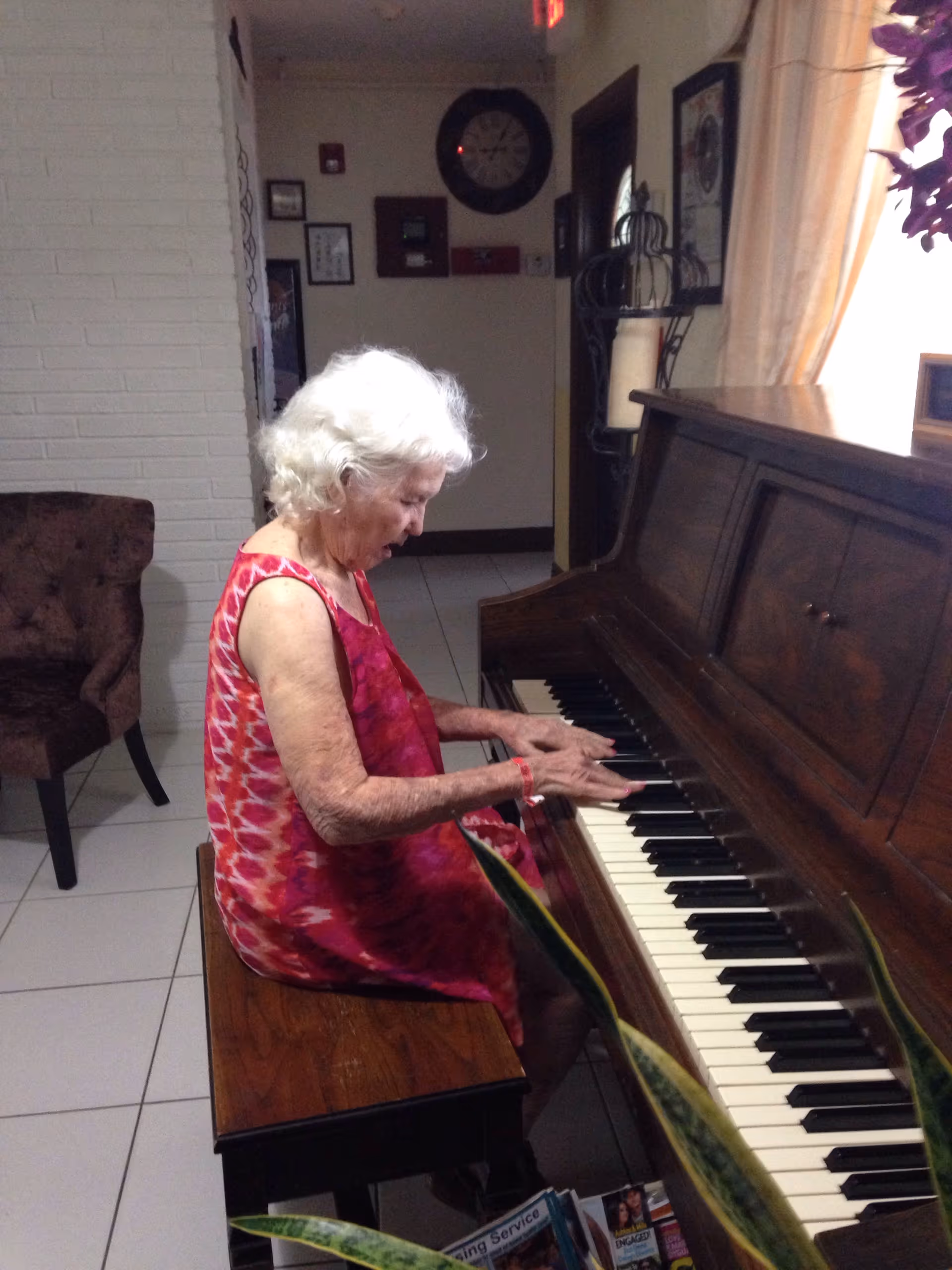 An elderly woman with white hair wearing a red patterned dress is sitting on a wooden bench playing a brown upright piano in a room with white tiled floor and light-colored walls. There is a brown upholstered chair to the left and a large clock on the wall in the background.