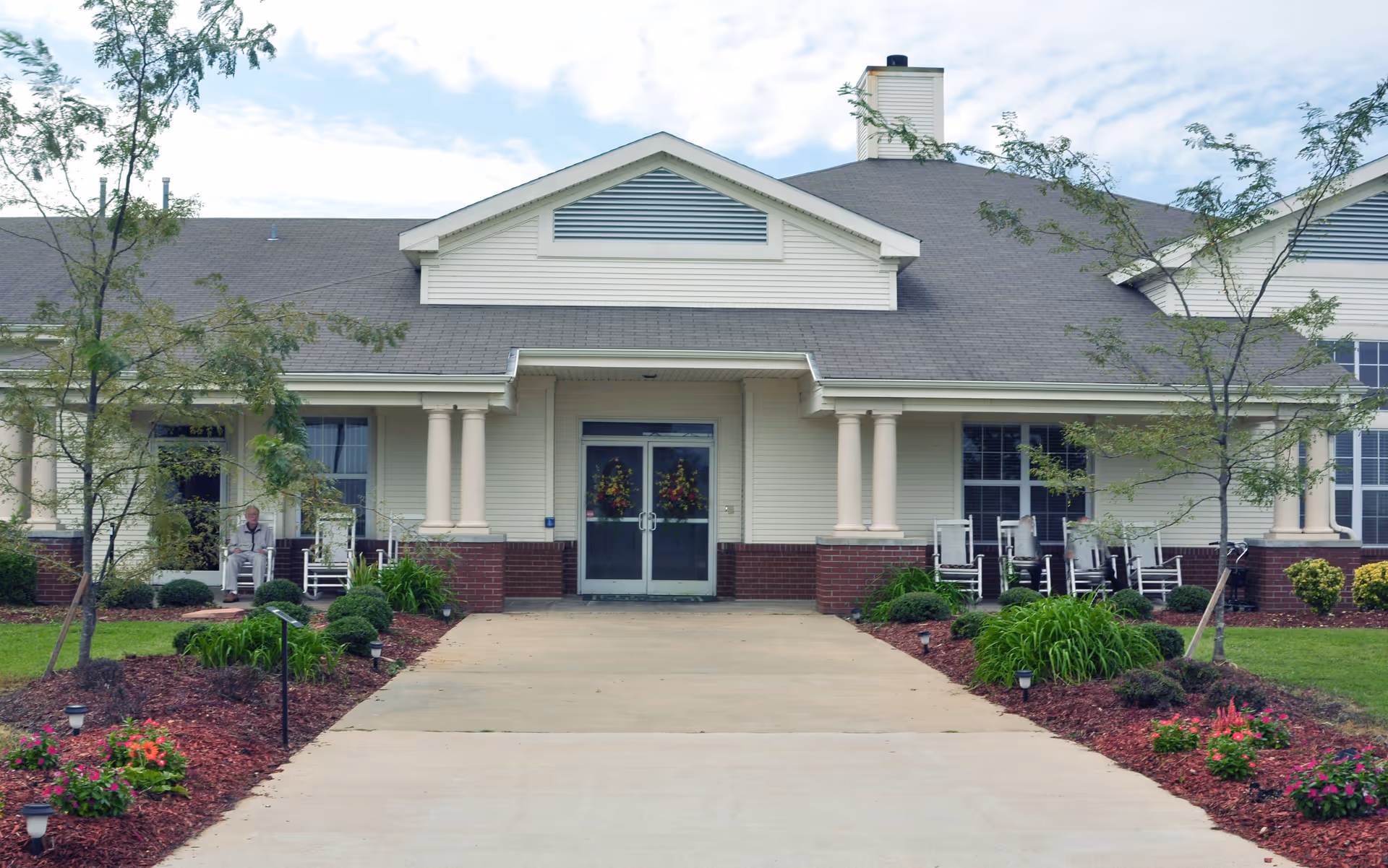 Front entrance of a single-story senior living building with double doors, columns, rocking chairs on the porch, and a landscaped walkway.