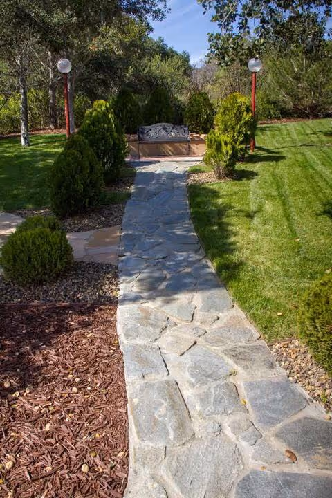 A stone pathway leading through a landscaped garden area with green grass, small bushes, and two lamp posts on either side. At the end of the path, there is a decorative bench surrounded by trees and shrubs under a clear blue sky.