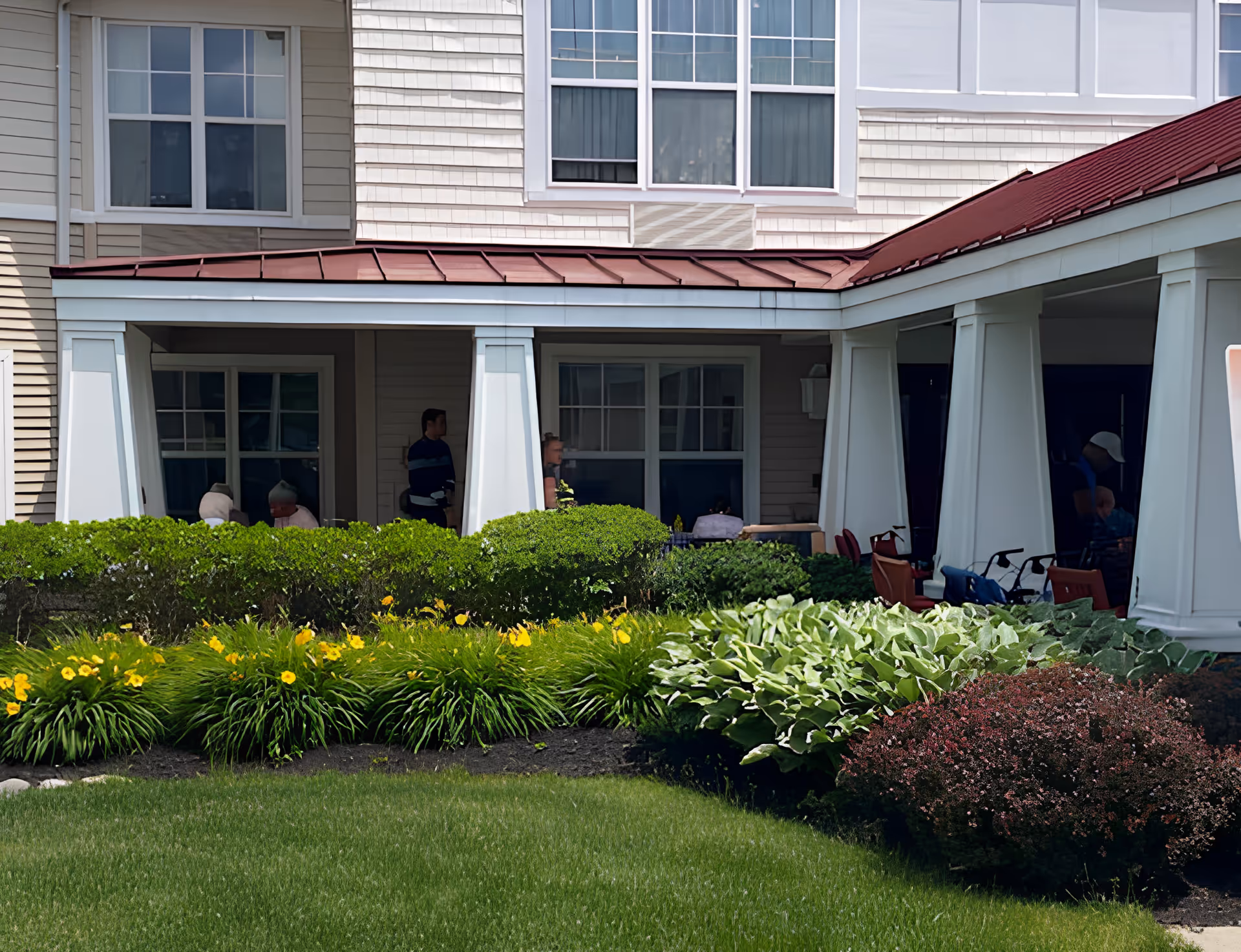 Covered front porch of a senior living building with white columns, landscaped flowerbeds and several people seated on the patio.