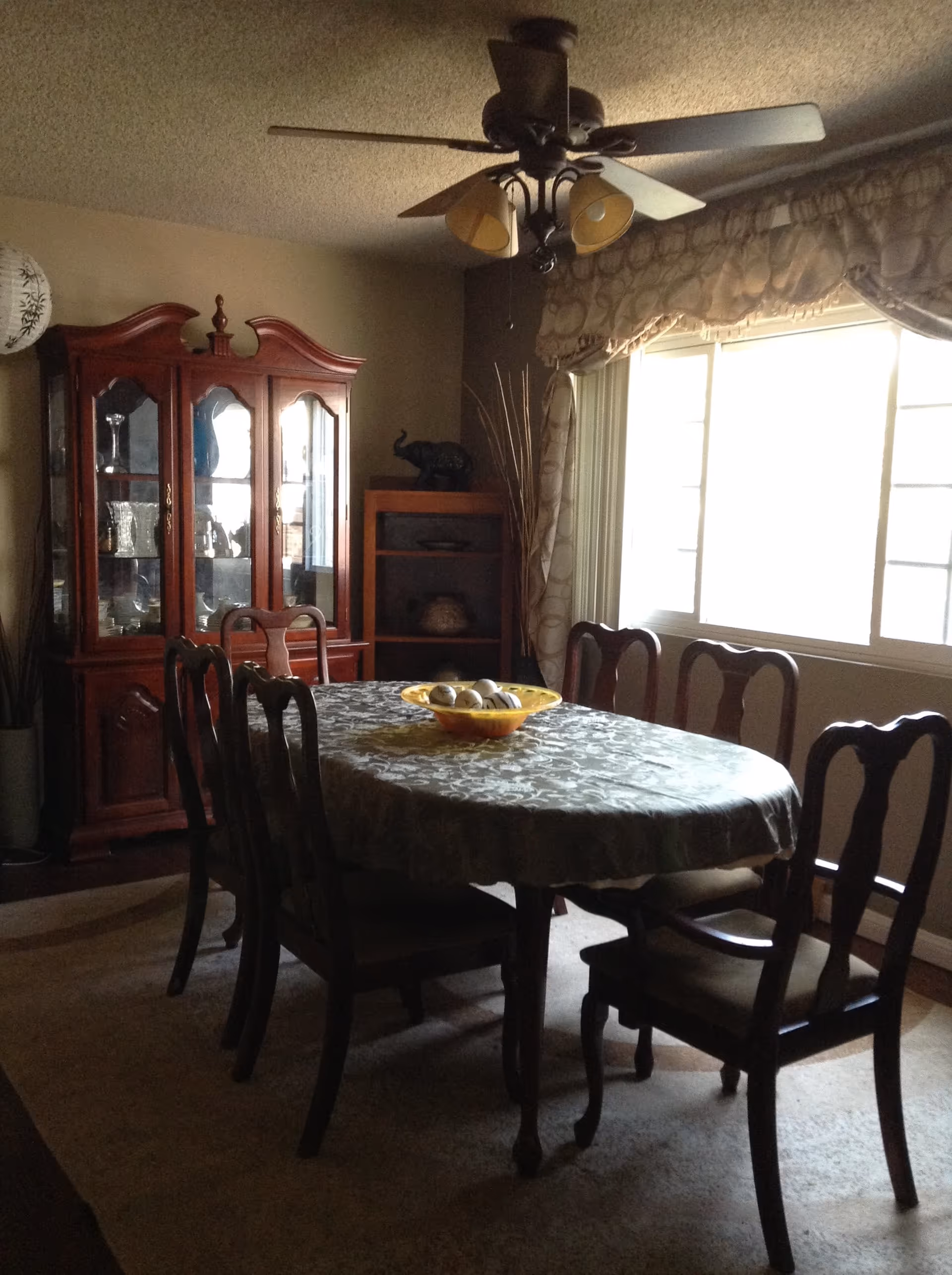 Dining room with an oval table and six chairs, a china cabinet, ceiling fan, and a large window.