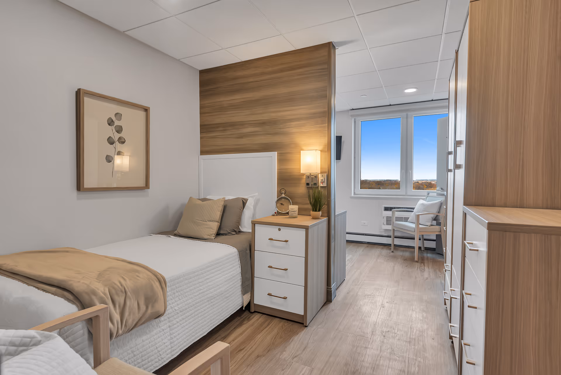 A bright and clean assisted living bedroom featuring a single bed with white and beige bedding, a wooden nightstand with drawers, a small clock, a plant, and a wall-mounted lamp. The room has light wood flooring, a large window with a view of the outdoors, a white chair with a cushion, and wooden cabinetry along the right wall. A framed botanical artwork hangs on the left wall.