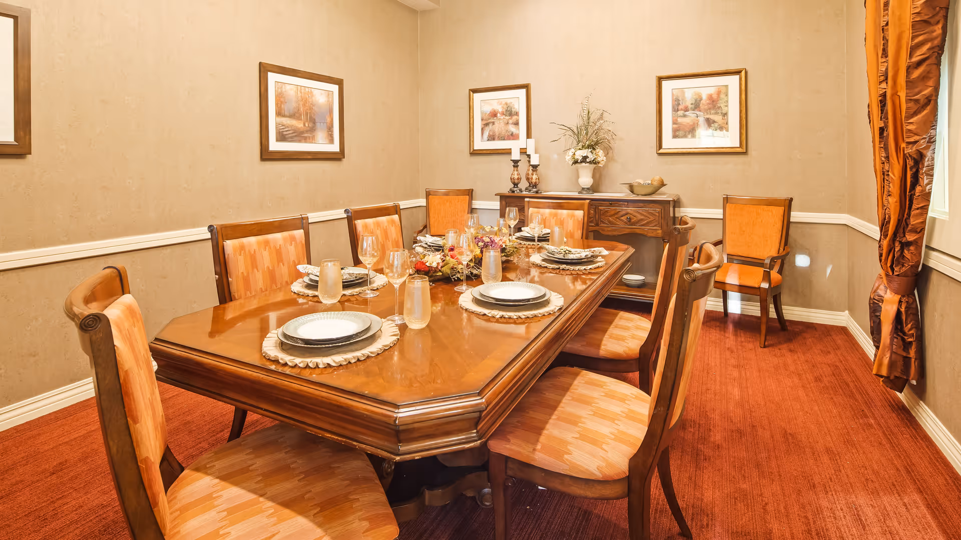 A formal dining room with a large wooden dining table set for six people. The table has plates, glasses, and decorative placemats. There are eight wooden chairs with orange patterned cushions around the table. The room has beige walls with three framed paintings and a sideboard with decorative items including candles and a flower arrangement. The floor is covered with a red carpet, and there is a window with orange curtains on the right side.