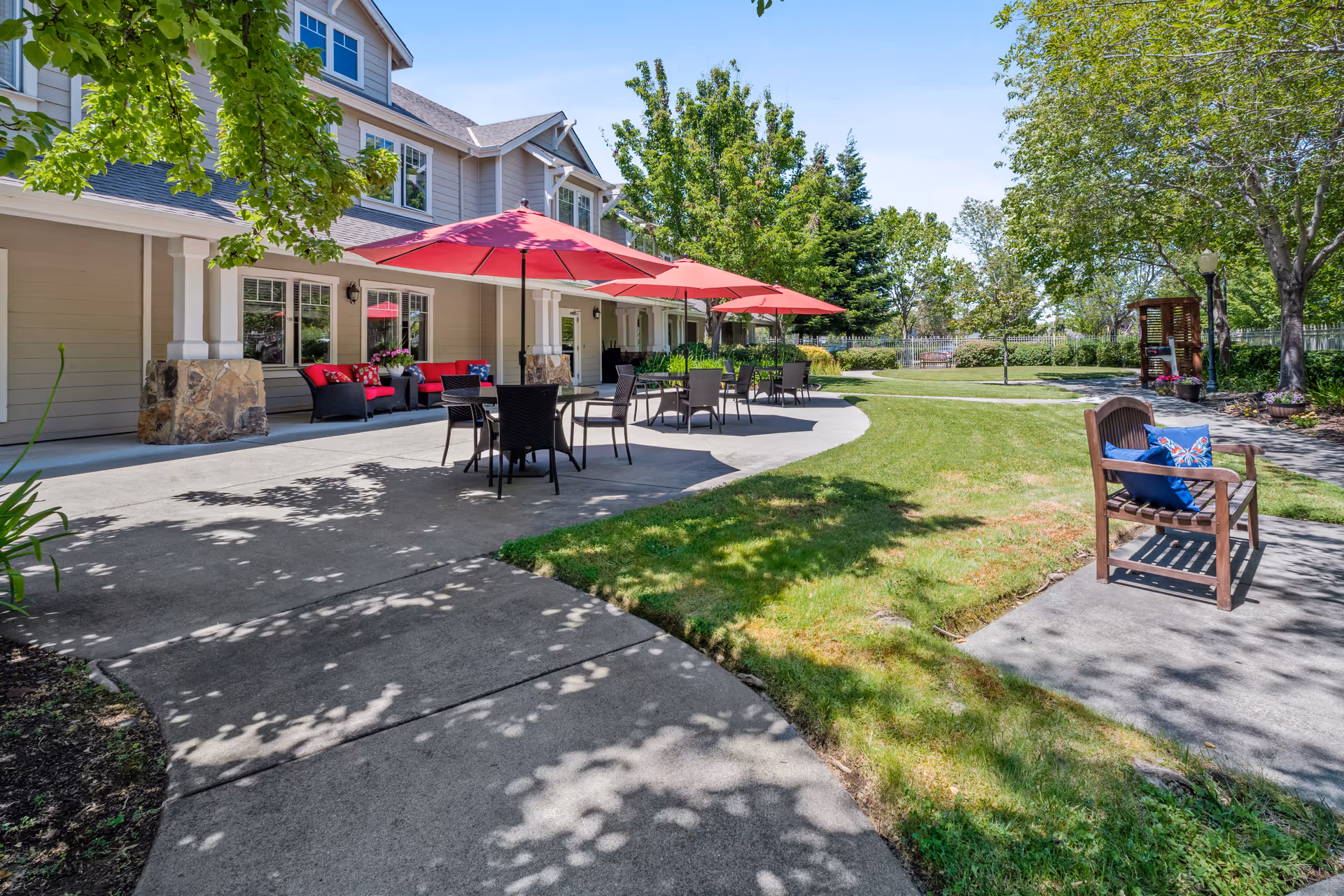 Outdoor patio area at Aegis Living Corte Madera with several tables and chairs under red umbrellas, a wooden bench with blue cushions on a concrete path, surrounded by green grass, trees, and shrubs on a sunny day.