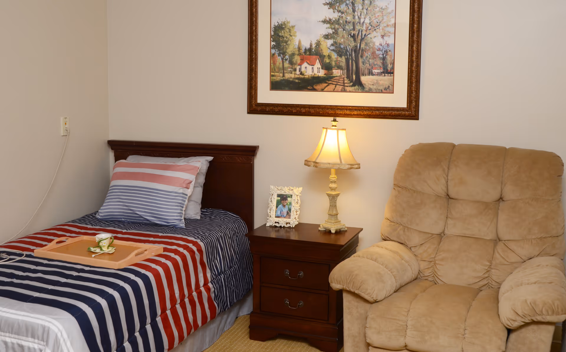A cozy bedroom with a single bed covered in a striped red, white, and blue comforter and matching pillow. A wooden nightstand beside the bed holds a decorative lamp and a framed photo. Next to the nightstand is a plush beige recliner chair. A framed painting of a countryside scene hangs on the wall above the nightstand.
