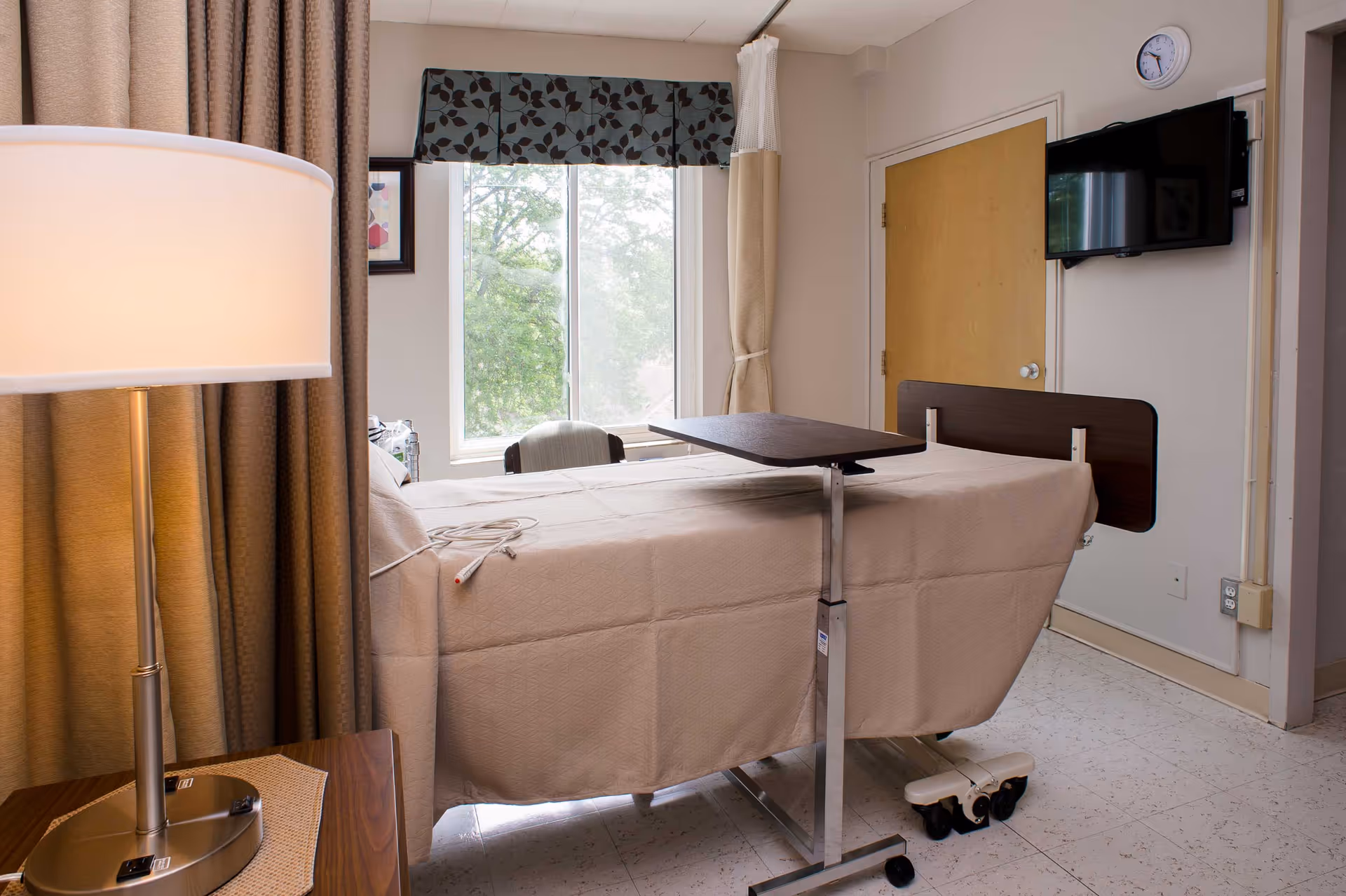 A hospital-style bed covered with a beige blanket in a room with a window showing green trees outside. Next to the bed is a small overbed table on wheels. A floor lamp with a white shade stands on a wooden side table with a woven placemat. The room has beige walls, a wooden door, and a wall-mounted flat-screen TV.