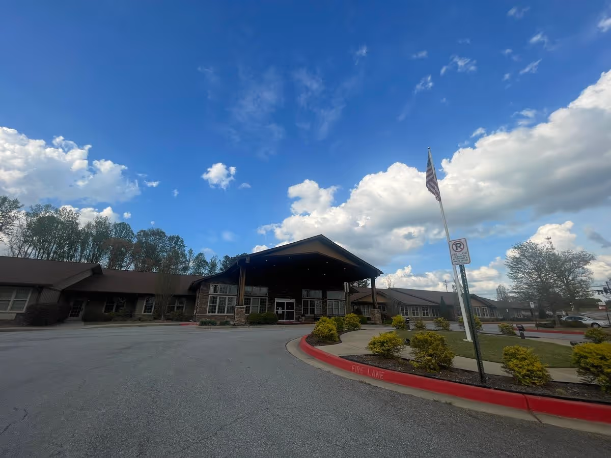 Front entrance of a senior living facility with a covered porte-cochère, flagpole, landscaped island, and driveway under a blue sky.