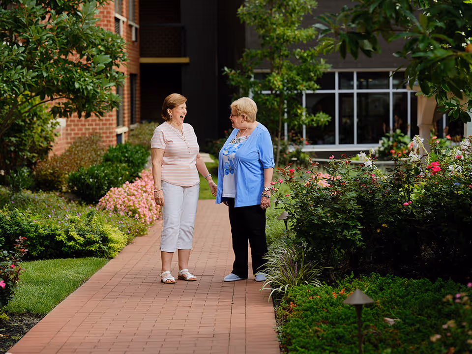 Two elderly women standing and talking on a brick pathway surrounded by lush green plants and colorful flowers, with a building visible in the background.