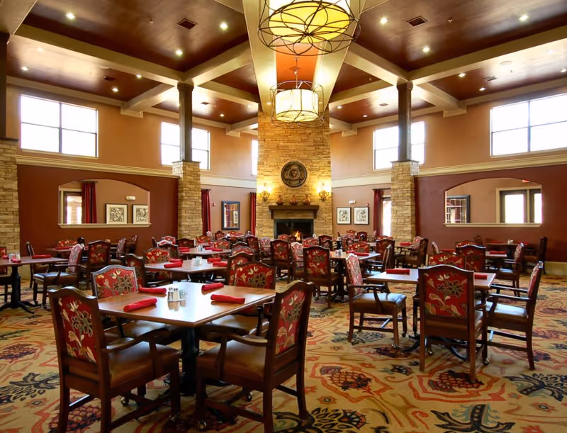 Spacious dining room with multiple wooden tables and chairs featuring floral upholstery. Each table is set with red napkins and salt and pepper shakers. The room has high ceilings with large windows letting in natural light, a stone fireplace in the center, and warm earth-tone walls and carpet with intricate patterns. Two large hanging light fixtures are visible from the ceiling.