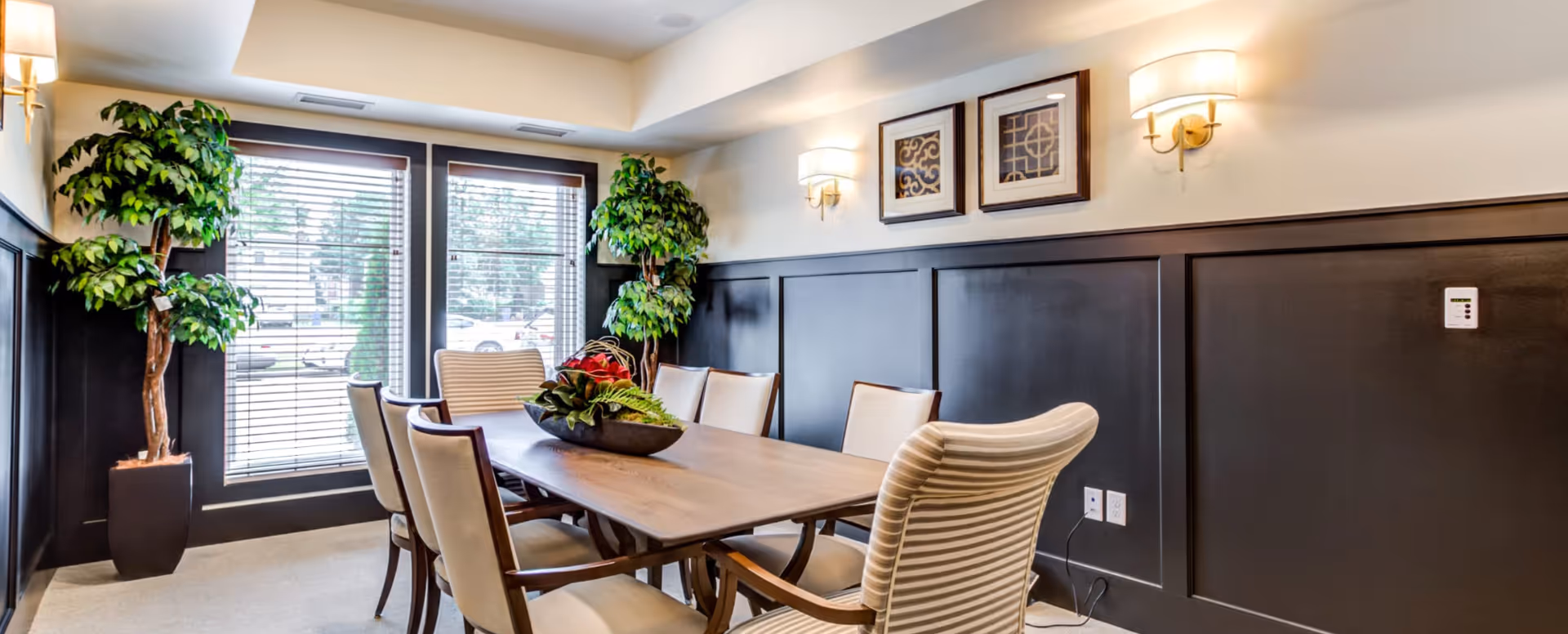 A well-lit dining room with a wooden table surrounded by six upholstered chairs. Two large potted plants are placed near the windows with blinds, and two framed artworks hang on the wall above dark wood paneling. Wall sconces provide additional lighting.